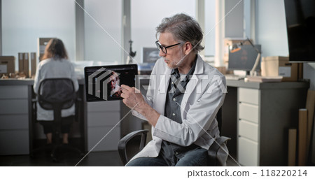 Mature scientist speaks, shows 3D model of human head on tablet during interview 118220214
