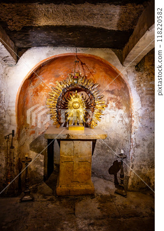 The crypt of the Basilica of Santa Maria in Cosmedin features a gilded altar adorned with sunburst motif under ancient stone arches, reflecting centuries of history in a serene, dimly lit environment. The crypt of the Basilica of Santa Maria in Cosmedin features a gilded altar adorned with sunburst motif under ancient stone arches, reflecting centuries of history in a serene, dimly lit environment. 118220582