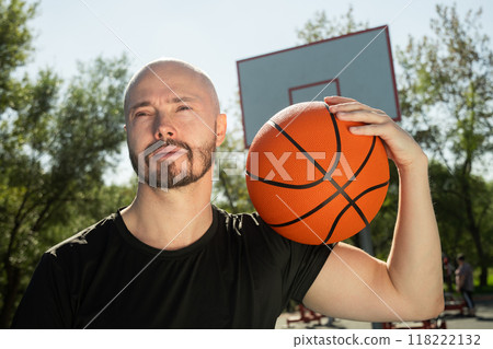 Happy basketball player holding basketball orange ball on shoulder on sports court. Basketball coach. Outdoors Happy basketball player holding basketball orange ball on shoulder on sports court. Basketball coach. Outdoors 118222132