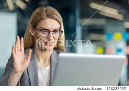Professional woman in office using laptop for video call. She waves hand at screen, engaged in virtual communication. Glasses and suit reflect work environment. Technology enhances teleconference. Professional woman in office using laptop for video call. She waves hand at screen, engaged in virtual communication. Glasses and suit reflect work environment. Technology enhances teleconference. 118222965