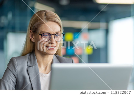 Businesswoman wearing glasses working on laptop in office. She appears confident and focused, surrounded by modern office interior. Professional attire suggests corporate environment 118222967