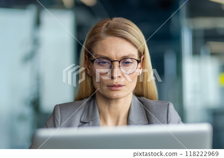 Professional businesswoman concentrating on computer screen in modern office. Wearing glasses and formal suit, she appears deep in thought, reflecting dedication and focus. Professional businesswoman concentrating on computer screen in modern office. Wearing glasses and formal suit, she appears deep in thought, reflecting dedication and focus. 118222969