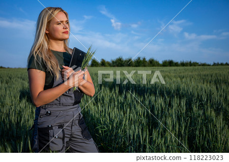 A female farmer standing in the wheat field, holding a tablet and wheat stalks 118223023