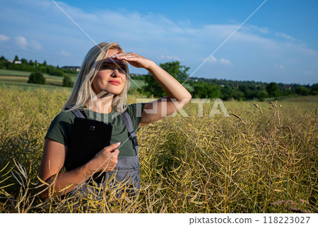 a woman farmer standing in the middle of rapeseed field, observing the horizon, shielding herself from the sun with hand and holding a tablet a woman farmer standing in the middle of rapeseed field, observing the horizon, shielding herself from the sun with hand and holding a tablet 118223027
