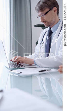 Male physician in white coat focused on laptop data in a modern clinic office while analyzing patient information on laptop. Medicine concept 118224016