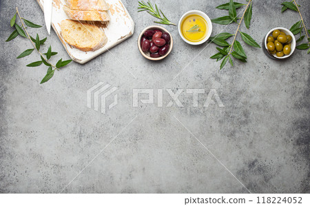 Composition with sliced fresh ciabatta on cutting board, green and brown olives, olive oil with rosemary, olive tree branches on gray concrete stone rustic background top view, copy space border Composition with sliced fresh ciabatta on cutting board, green and brown olives, olive oil with rosemary, olive tree branches on gray concrete stone rustic background top view, copy space border 118224052