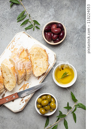 Sliced fresh ciabatta on cutting kitchen board, green and brown olives, olive oil with rosemary, olive tree branches on gray concrete stone rustic background top view copy space 118224053