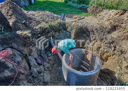 Installing septic tank from concrete rings with your own hands, homeowner connects sewer pipe to hole in cement wall of well. 118225935