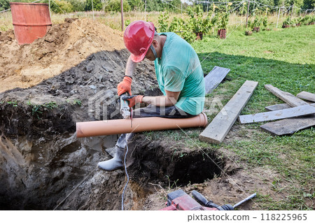 Worker uses an abrasive wheel on an angle grinder to cut off section of plastic sewer pipe. 118225965