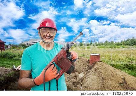 Mature smiling middle aged man in construction helmet and safety glasses holds corded hammer drill with chisel in his hands. 118225970