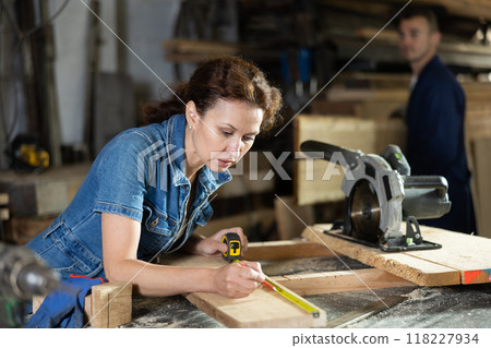 Woman employee of carpentry workshop makes measurements of wooden board, counts off set parameters Woman employee of carpentry workshop makes measurements of wooden board, counts off set parameters 118227934
