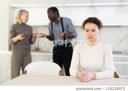 Middle-aged woman sitting at the kitchen table with her back to man and old woman quarreling together Middle-aged woman sitting at the kitchen table with her back to man and old woman quarreling together 118228073