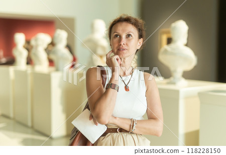 Interested woman looking closely at classical statues in museum and holding tour sheet in hands 118228150