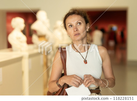 Focused pretty woman visitor with booklet in her hands examines exhibit standing and exploring old statues in museum 118228151