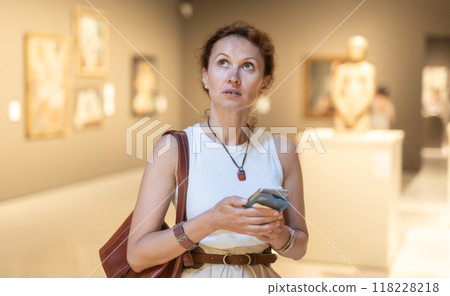 Attractive young woman in casual clothing with phone in hands looking painting in museum 118228218