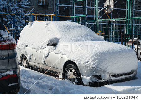 A snow-covered car is parked in the parking lot. The tires, wheels and hood of the car are covered with a white coating. 118231047