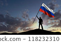 The silhouette of a man, standing on a small mountain, waves the Russian flag, in the background a cloudy sky at dawn 118232310