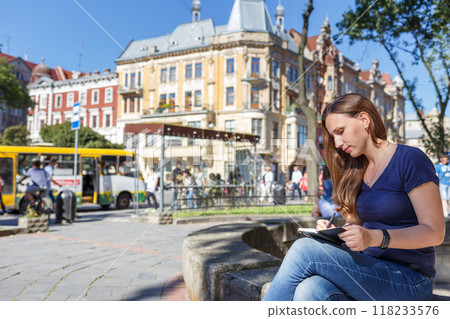 Young woman writing in a notebook on a stone bench in the city 118233576