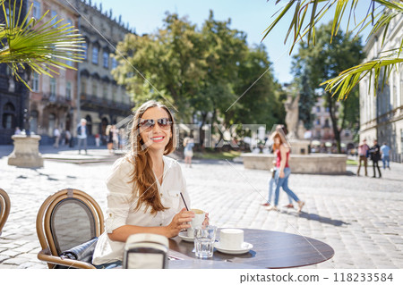 Young woman enjoying a sunny afternoon at an outdoor cafe in a charming European square 118233584