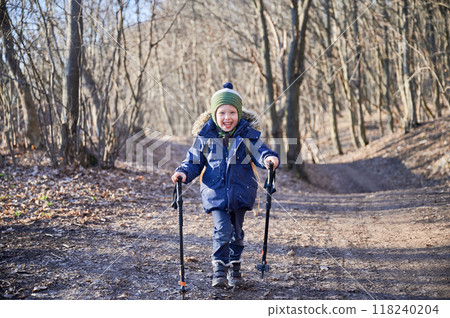 Young child smiling while hiking on forest trail, bundled up in blue winter jacket with fur-lined hood and green striped beanie. Kid holding two trekking poles and enjoying sunny day outdoors. 118240204