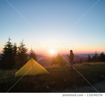 Tranquil sunrise over mountain campsite, with vibrant yellow tent and lone camper standing nearby. Tall evergreens silhouetted against soft morning light, with rolling hills fading into horizon. Tranquil sunrise over mountain campsite, with vibrant yellow tent and lone camper standing nearby. Tall evergreens silhouetted against soft morning light, with rolling hills fading into horizon. 118240256