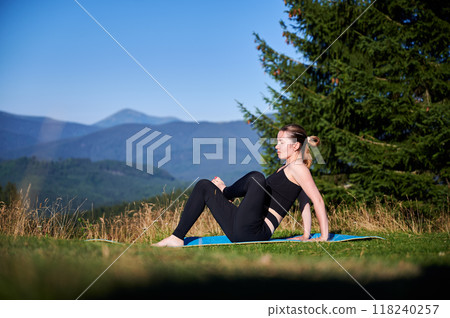 Woman practicing yoga outdoors in the mountains in a serene, natural setting. Female performing yoga pose on mat, with backdrop of beautiful mountain landscape at sunrise or sunset. 118240257