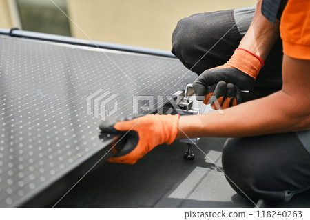Worker building photovoltaic solar panel system on rooftop of house. Close up of man engineer in gloves installing solar module with help of hex key outdoors. Renewable energy generation concept. 118240263