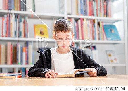 11-year-old teenage boy in a white T-shirt reads an interesting book in the library against the background of bookshelves 11-year-old teenage boy in a white T-shirt reads an interesting book in the library against the background of bookshelves 118240406