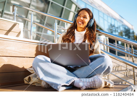 Young woman in headphones with laptop on city street. Video conference. ?nline training. Freelancer 118240484