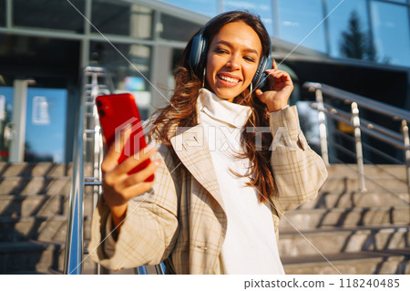 Beautiful woman using mobile phone in city while listening music through earphones. Music, emotions. 118240485