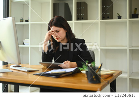 Exhausted young businesswoman sitting at office desk suffering from headache 118240615
