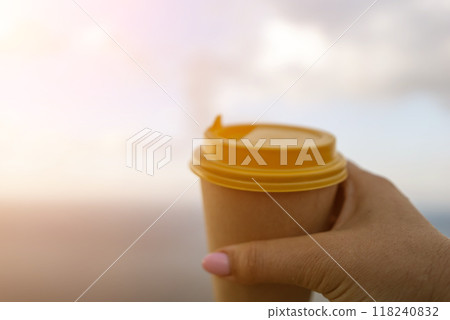 Hand holding Yellow cup with lid, coffee against a backdrop of a blue sky and sea. Illustrating cup and beverage Hand holding Yellow cup with lid, coffee against a backdrop of a blue sky and sea. Illustrating cup and beverage 118240832