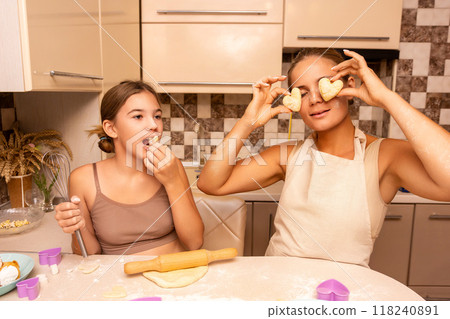 Mother Daughter Kitchen Fun Cooking - Two women, a mother and daughter, are cooking in a kitchen. The daughter is holding a rolling pin and the mother is holding two pieces of dough. They are both 118240891