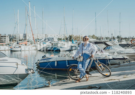 Woman enjoys bike ride along waterfront, Marina surroundings. She is wearing a white shirt and blue jeans, and she has a handbag with her. Capturing outdoor bike ride by waterfront. Woman enjoys bike ride along waterfront, Marina surroundings. She is wearing a white shirt and blue jeans, and she has a handbag with her. Capturing outdoor bike ride by waterfront. 118240893