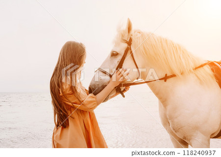 A white horse and a woman in a dress stand on a beach, with the 118240937