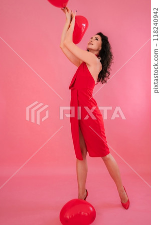 A woman in a red dress is posing with two red balloons. The balloons are on the floor, and the woman is standing in front of them. Concept of playfulness and fun. A woman in a red dress is posing with two red balloons. The balloons are on the floor, and the woman is standing in front of them. Concept of playfulness and fun. 118240942