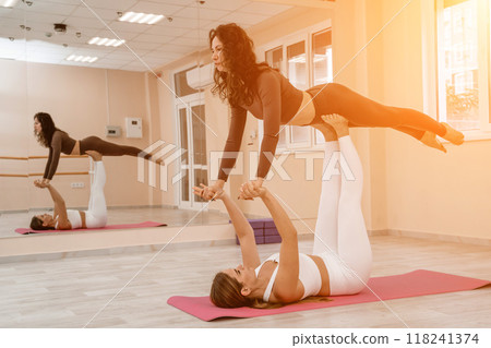 Two women do acroyoga on a light floor near a large mirror in a yoga studio. They are wearing leggings and tops. Two women do acroyoga on a light floor near a large mirror in a yoga studio. They are wearing leggings and tops. 118241374