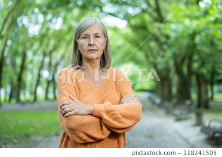 Mature woman with gray hair in orange sweater stands confidently with arms crossed in serene park setting. Her expression conveys strength, resilience, and contemplation. Mature woman with gray hair in orange sweater stands confidently with arms crossed in serene park setting. Her expression conveys strength, resilience, and contemplation. 118241505