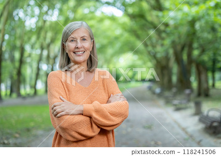Mature woman with gray hair and orange sweater stands confidently in lush green park. She smiles warmly, symbolizing happiness, serenity, and appreciation for nature. Mature woman with gray hair and orange sweater stands confidently in lush green park. She smiles warmly, symbolizing happiness, serenity, and appreciation for nature. 118241506