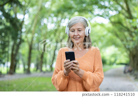 Senior woman wearing headphones, using phone outdoors in park setting. She appears happy, engaged with technology, enjoying music. Represents connection, technology usage among older generation 118241510