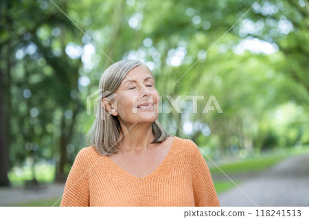 Elderly woman with short gray hair wearing orange sweater stands with eyes closed, enjoying tranquility in green park. Represents relaxation, mindfulness, and connection with nature. Elderly woman with short gray hair wearing orange sweater stands with eyes closed, enjoying tranquility in green park. Represents relaxation, mindfulness, and connection with nature. 118241513