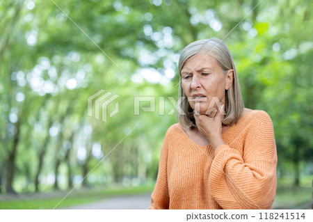 Mature woman with gray hair experiencing sore throat while outdoors among green trees. Wearing orange sweater, she appears uncomfortable, touching her throat. 118241514