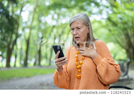 Elderly woman looking at smartphone with concern and disbelief while outdoors. Expresses emotions of worry and surprise, reacting to unexpected news. 118241523