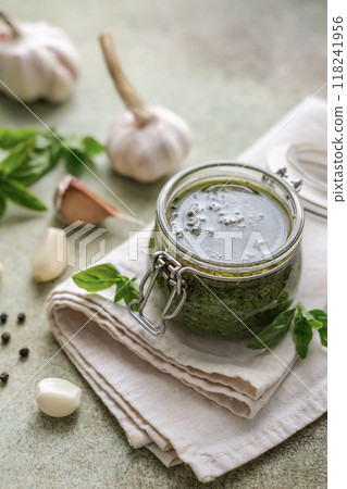 Homemade pesto sauce in a small glass jar, basil, garlic, pine nuts and black pepper on the table 118241956