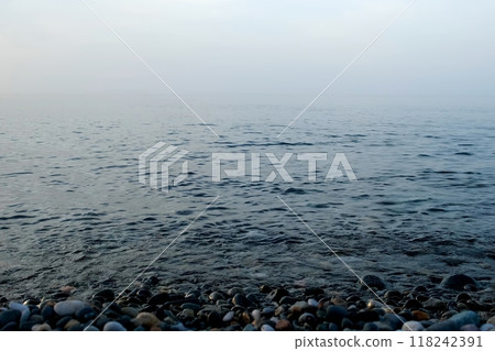 Pebble beach with small pebbles calm sea water surface and fog over sea. Pebble beach with small pebbles calm sea water surface and fog over sea. 118242391