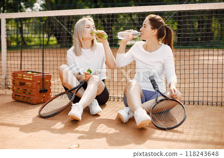 Two young women sitting by tennis net. Having conversation after game.Summer, sports, leisure. 118243648