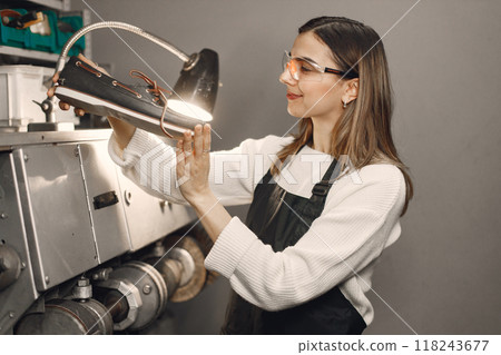 Young woman working in her workspace. Woman wearing an apron and repairing a shoes. Woman looking on a shoe in an eyeglasses under a lamp. 118243677