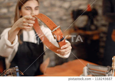 Young woman working in her workspace. Woman wearing an apron and making a belt. Grounge dark stone texture background. Young woman working in her workspace. Woman wearing an apron and making a belt. Grounge dark stone texture background. 118243687