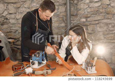 Mature craftsman with young woman working in workspace. Man and woman wearing an apron and making a belt. Grounge dark stone texture background. Mature craftsman with young woman working in workspace. Man and woman wearing an apron and making a belt. Grounge dark stone texture background. 118243699