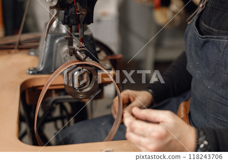 Cropped photo of mature craftsman working in his workspace. Man wearing an apron and using a sewing machine. Grounge dark stone texture background. Cropped photo of mature craftsman working in his workspace. Man wearing an apron and using a sewing machine. Grounge dark stone texture background. 118243706